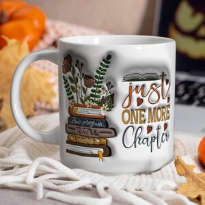 Pode incluir: Caneca de cerâmica branca com um design de livros empilhados, flores e o texto "Just One More Chapter". A caneca está sobre uma superfície branca e felpuda com folhas de outono e abóboras.