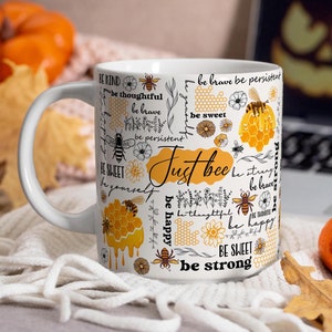 Puede incluir: Taza de cerámica blanca con un diseño de abeja amarilla y negra. La taza presenta el texto "Just bee" y varias afirmaciones positivas, como "sé amable", "sé valiente" y "sé feliz".