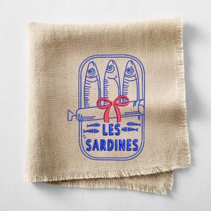 Peut inclure: Une serviette carr&eacute;e en lin beige aux bords effiloch&eacute;s pr&eacute;sentant une broderie de trois sardines dans une bo&icirc;te. Le contour bleu repr&eacute;sente les poissons et le contenant, agr&eacute;ment&eacute; d'un n&oelig;ud rouge et du texte bleu LES SARDINES centr&eacute;.