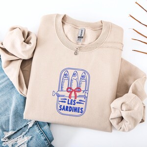Peut inclure: Un sweat-shirt &agrave; col rond beige avec un motif repr&eacute;sentant trois sardines dans une bo&icirc;te de conserve ouverte. Le design inclut un n&oelig;ud rouge, de petites ic&ocirc;nes de poissons et le texte bleu "LES SARDINES" imprim&eacute; sous la bo&icirc;te. &Eacute;tiquette Gildan.