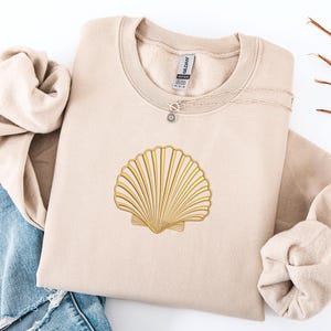 May include: A beige sweatshirt with a gold seashell design. A silver necklace with a sun pendant is on the sweatshirt. The sweatshirt is folded with a pair of blue jeans on the left side.