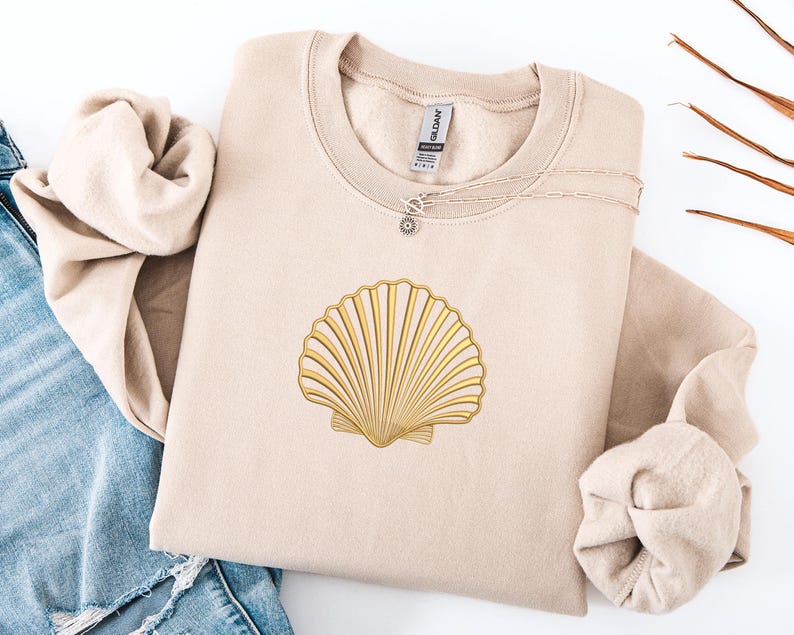 May include: A beige sweatshirt with a gold seashell design. A silver necklace with a sun pendant is on the sweatshirt. The sweatshirt is folded with a pair of blue jeans on the left side.