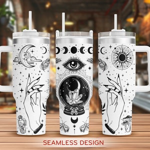 May include: A white stainless steel tumbler with a black and white design featuring a hand, a crystal ball, a crescent moon, stars, an eye, and a sun. The tumbler has a lid and a handle.