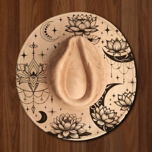 Può includere: Un cappello fedora beige con tesa larga. La tesa è decorata con disegni floreali e celesti neri, tra cui fiori di loto, lune crescenti e stelle. Il cappello ha una corona a forma di goccia.