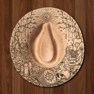 May include: A tan fedora hat featuring a detailed black line art print across the brim. The design includes illustrations of a skull, tarot cards, candles, potion bottles, crystals, and celestial motifs like a crescent moon and stars.