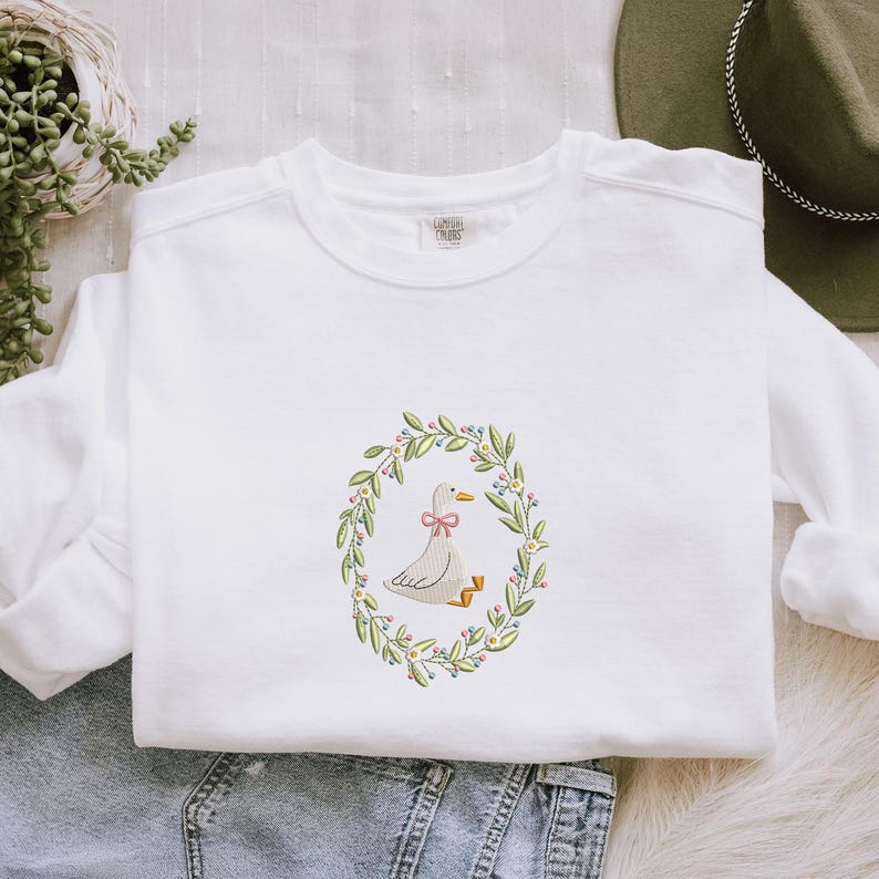 Puede incluir: Sudadera blanca con un dise&ntilde;o bordado de un pato con un lazo rosa, rodeado por una corona floral. El dise&ntilde;o est&aacute; centrado en la parte delantera. La sudadera est&aacute; hecha de un material suave.