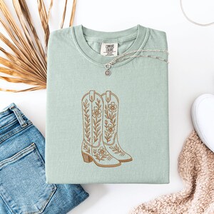 May include: A light green Comfort Colors t-shirt with an embroidered design of two brown cowboy boots with floral details. A silver necklace with a small pendant rests on the shirt. The shirt is paired with blue jeans and a white shoe.