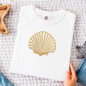May include: White t-shirt featuring a gold seashell design. The seashell is centered on the shirt and has detailed lines. The shirt is folded and placed on a textured surface. A hand is holding the shirt. A wooden star-shaped tray and a star-shaped decoration are also visible.