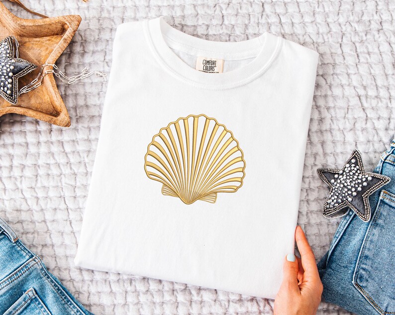 May include: White t-shirt featuring a gold seashell design. The seashell is centered on the shirt and has detailed lines. The shirt is folded and placed on a textured surface. A hand is holding the shirt. A wooden star-shaped tray and a star-shaped decoration are also visible.