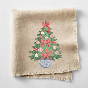 May include: A square beige linen napkin with a frayed edge featuring an embroidered green Christmas tree. The tree is decorated with red bows and round ornaments in shades of red and white, and it sits in a blue and white floral patterned pot.
