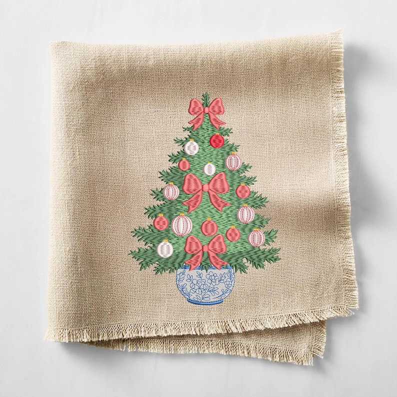 May include: A square beige linen napkin with a frayed edge featuring an embroidered green Christmas tree. The tree is decorated with red bows and round ornaments in shades of red and white, and it sits in a blue and white floral patterned pot.