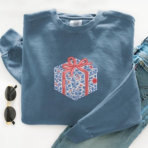 May include: A blue crewneck sweatshirt featuring an embroidered graphic of a gift box. The box is decorated with a blue and white floral pattern, red berries, and a large pink ribbon bow. The interior neck label reads Comfort Colors.