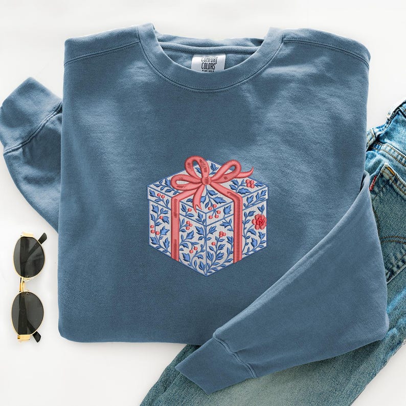 May include: A blue crewneck sweatshirt featuring an embroidered graphic of a gift box. The box is decorated with a blue and white floral pattern, red berries, and a large pink ribbon bow. The interior neck label reads Comfort Colors.