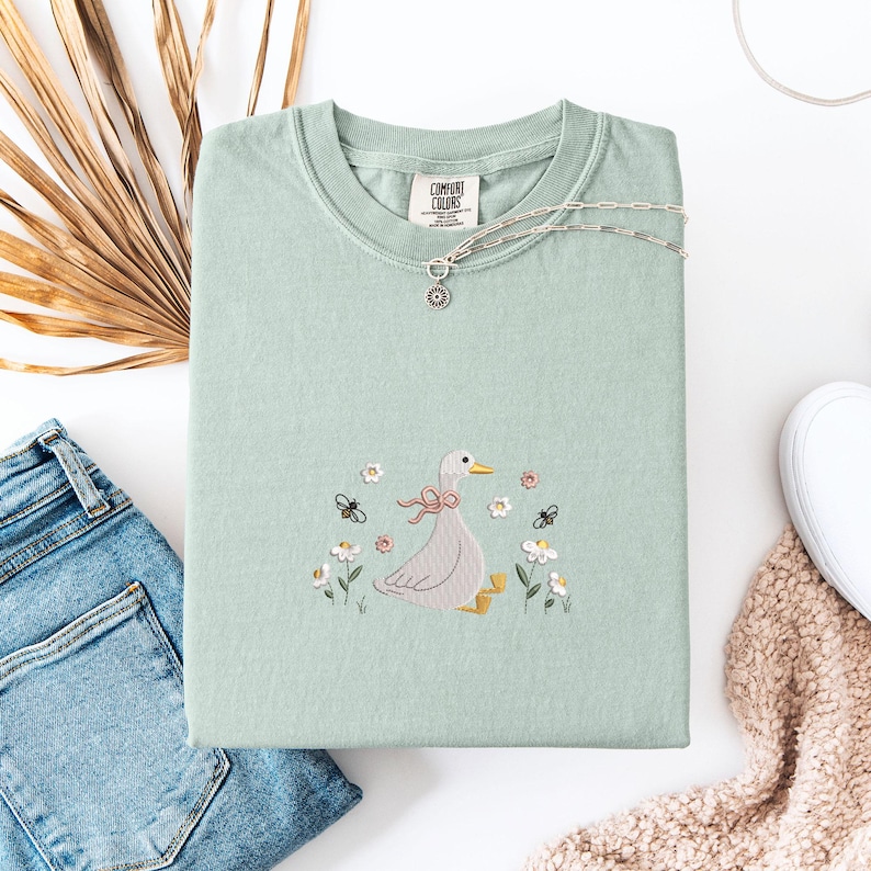Op de afbeelding: Een lichtgroene Comfort Colors sweatshirt met een borduursel van een gans met een roze strik, bloemen en vlinders. Een zilveren ketting met een hanger ligt op de trui. Een blauwe jeans en een witte schoen zijn ook zichtbaar.