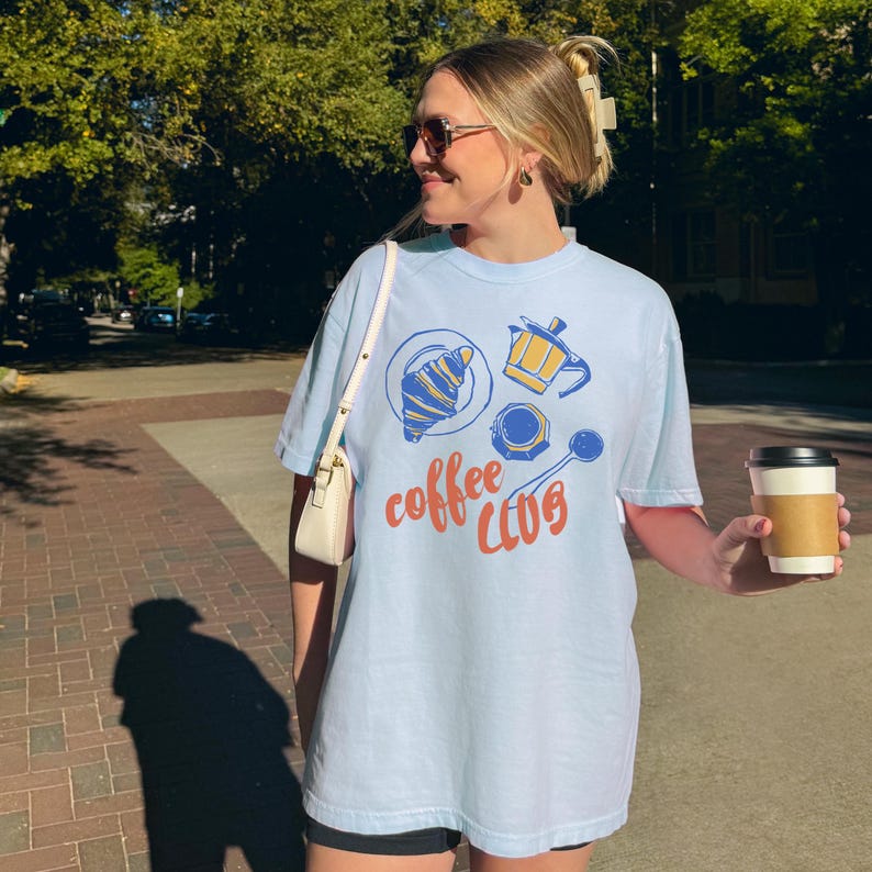 May include: Pale blue t-shirt featuring a coffee-themed design. The graphic includes a croissant, coffee pot, coffee cup, and spoon. The words "coffee CLUB" are written in orange script. A person is holding a takeaway coffee cup.