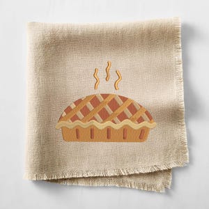 May include: A beige linen napkin with a fringed edge, embroidered with a pie design. The pie has a lattice crust and steam rising from it, suggesting it's freshly baked. A cozy kitchen or dining accessory.