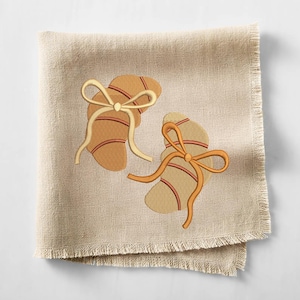 May include: A beige linen napkin with a fringed edge. Two embroidered designs of crescent-shaped pastries with red stripes and cream-colored bows are featured on the napkin. The design is a warm, inviting style.