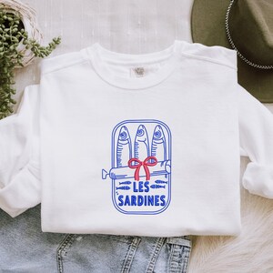 Peut inclure: Un sweat-shirt blanc &agrave; manches longues avec une broderie bleue repr&eacute;sentant trois sardines dans une bo&icirc;te de conserve ouverte. Un petit n&oelig;ud rouge est nou&eacute; au centre de la bo&icirc;te. Le texte LES SARDINES est brod&eacute; en bleu sous le motif.