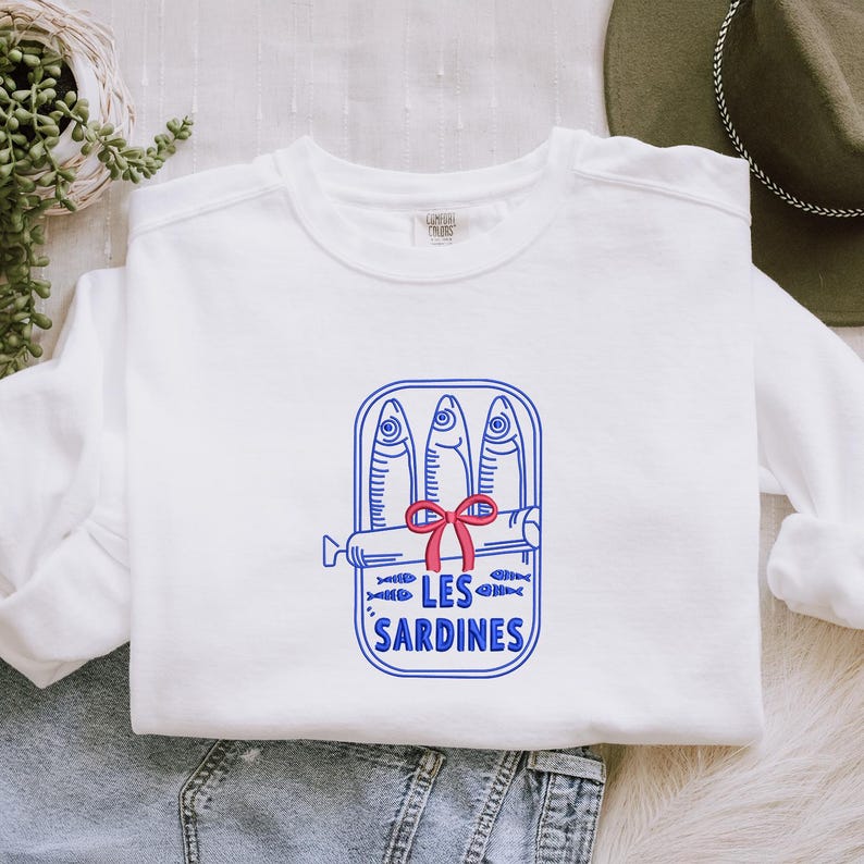 Peut inclure: Un sweat-shirt blanc &agrave; manches longues avec une broderie bleue repr&eacute;sentant trois sardines dans une bo&icirc;te de conserve ouverte. Un petit n&oelig;ud rouge est nou&eacute; au centre de la bo&icirc;te. Le texte LES SARDINES est brod&eacute; en bleu sous le motif.