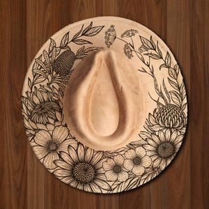 Puede incluir: Sombrero fedora beige con diseño floral. El sombrero presenta una detallada ilustración botánica en blanco y negro alrededor del ala, incluyendo girasoles y otras flores. El sombrero tiene una corona en forma de lágrima.
