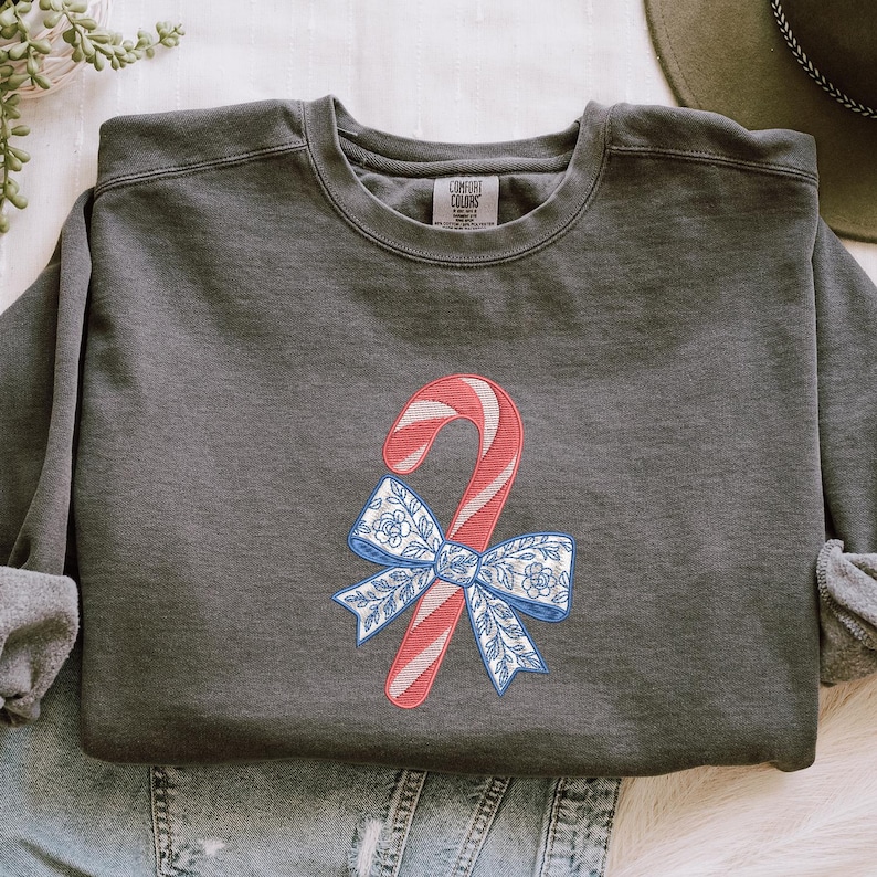 May include: A charcoal gray crewneck sweatshirt featuring an embroidered design of a red and white striped candy cane tied with a blue and white floral patterned bow. The garment has a Comfort Colors brand label visible at the inner neckline.