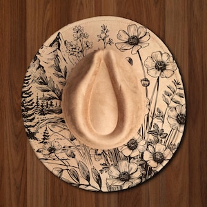 May include: A beige fedora hat with a floral and mountain landscape design. The brim features black ink illustrations of flowers, trees, and a mountain range. The hat has a teardrop-shaped crown.
