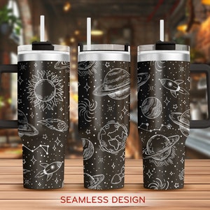 May include: Three black insulated tumblers with white line art of planets, stars, rockets, and a sun. Each tumbler has a black handle, a silver rim, and a white straw. The words "SEAMLESS DESIGN" are at the bottom.