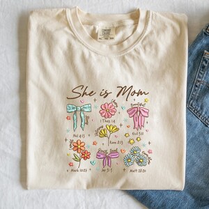 May include: Cream-colored t-shirt with the text "She is Mom" and floral and bow graphics. The design includes Bible verses. The shirt is folded next to a pair of blue jeans.