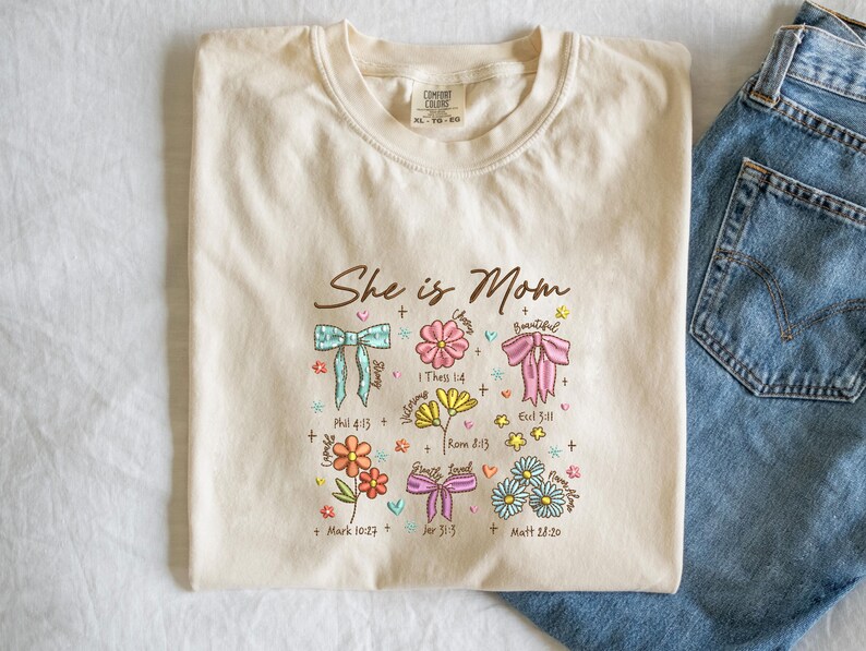 May include: Cream-colored t-shirt with the text "She is Mom" and floral and bow graphics. The design includes Bible verses. The shirt is folded next to a pair of blue jeans.