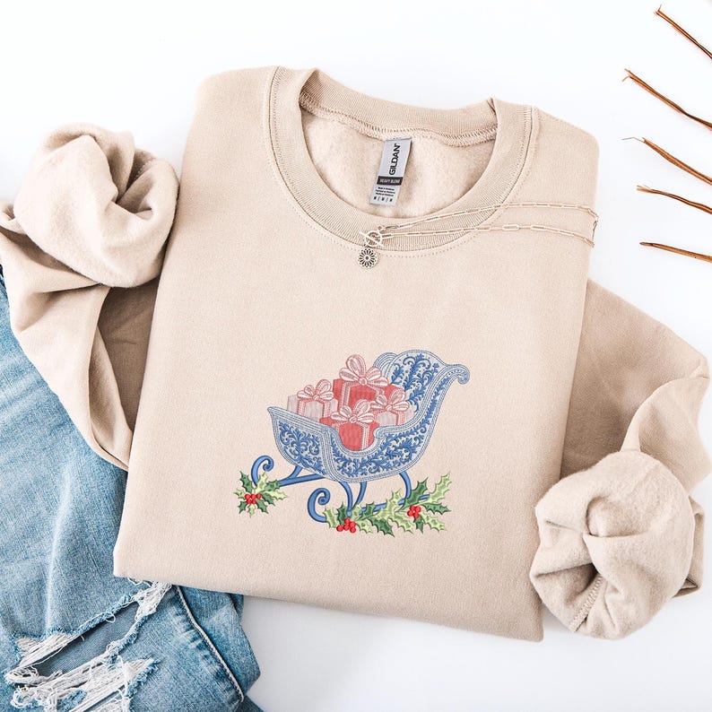 May include: A beige crewneck sweatshirt featuring an embroidered design of a blue sleigh filled with red gift boxes and topped with white bows. The embroidery includes green holly leaves and red berries at the base of the sleigh. A silver chain necklace rests on the collar.