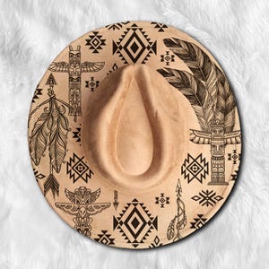 Puede incluir: Un sombrero fedora de color tostado con un patrón impreso en negro de tótems, plumas, flechas y formas geométricas de diamante. El sombrero tiene una corona pellizcada y un ala ancha, con dibujos detallados de líneas negras en la superficie.