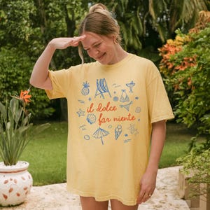 Puede incluir: Una camiseta de manga corta de color amarillo p&aacute;lido con un estampado gr&aacute;fico central en azul y naranja. El dise&ntilde;o incluye el texto "il dolce far niente" rodeado de ilustraciones de una silla de playa, pi&ntilde;a, bikini, concha, sombrilla y helado.