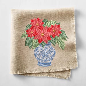 May include: A square beige linen napkin with frayed edges featuring an embroidered design of red poinsettia flowers with green leaves arranged in a blue and white patterned vase. The embroidery displays detailed floral motifs and textures.