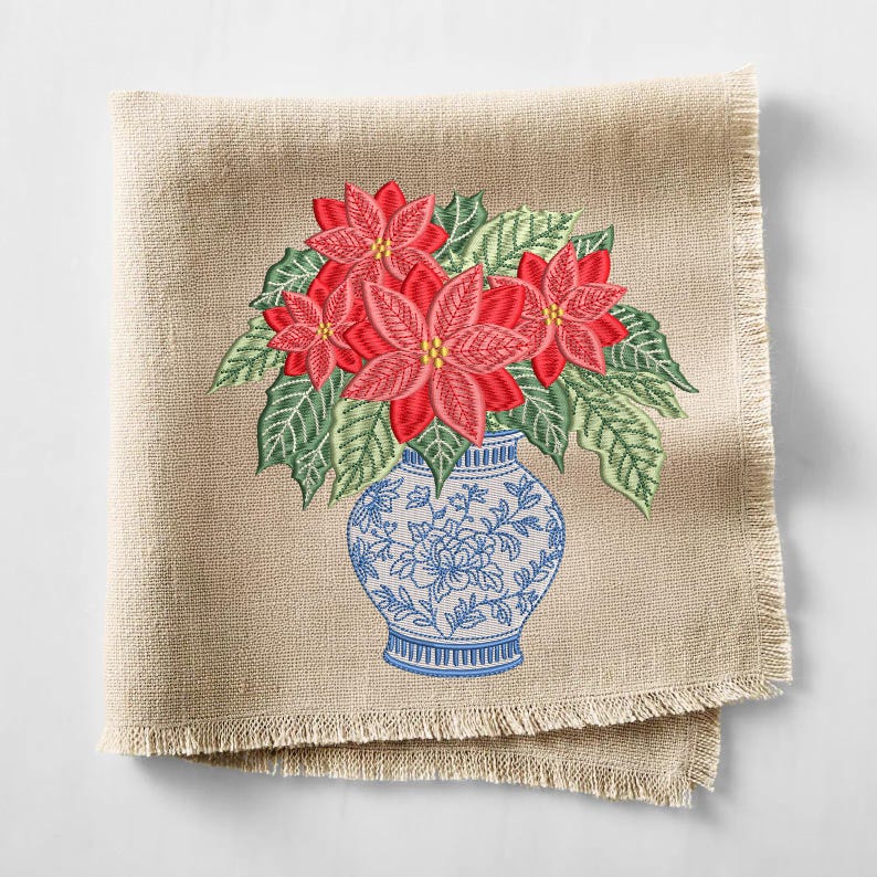 May include: A square beige linen napkin with frayed edges featuring an embroidered design of red poinsettia flowers with green leaves arranged in a blue and white patterned vase. The embroidery displays detailed floral motifs and textures.