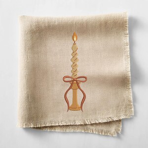 May include: A beige linen napkin with a fringed edge. An embroidered candle design features a twisted candle with a flame, a bow, and a candle holder in shades of brown and orange.