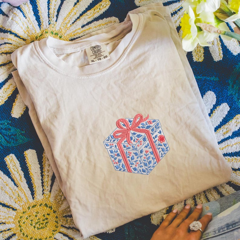 May include: A beige short-sleeve t-shirt featuring an embroidered graphic of a gift box with a red ribbon. The box has a blue floral and berry pattern. The shirt has a Comfort Colors brand label at the neck with the size marked as XL-TG-EG.