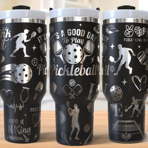 May include: A black stainless steel tumbler with a silver design featuring a pickleball paddle and ball, the text "It's a good day to play pickleball", and other pickleball-themed graphics.