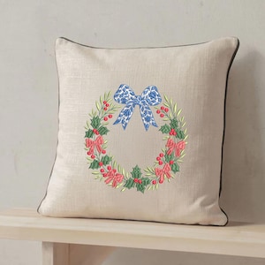 May include: A square beige throw pillow with a dark brown piped edge. The center features an embroidered wreath of green holly leaves and red berries, accented with small pink bows and a large blue patterned bow at the top of the wreath.