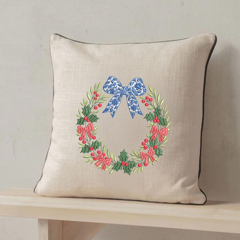 May include: A square beige throw pillow with a dark brown piped edge. The center features an embroidered wreath of green holly leaves and red berries, accented with small pink bows and a large blue patterned bow at the top of the wreath.