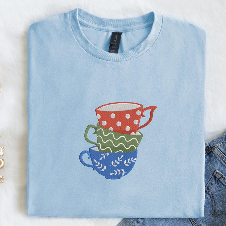 May include: A light blue t-shirt featuring an embroidered design of three stacked teacups. The top cup is red with white polka dots, the middle is green with a wavy pattern, and the bottom is blue with leaf designs.