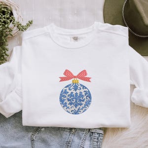 May include: A white crewneck sweatshirt featuring an embroidered design of a round Christmas ornament. The ornament has a blue and white floral pattern with a gold cap and a red and white striped bow tied at the top of the ornament.