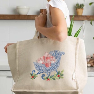 May include: A beige jute tote bag featuring an embroidered design of a blue sleigh filled with red and white striped gift boxes. The sleigh is decorated with intricate blue scrollwork patterns and accented with green holly leaves and red berries.
