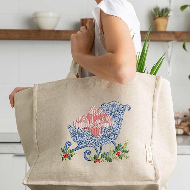 May include: A beige jute tote bag featuring an embroidered design of a blue sleigh filled with red and white striped gift boxes. The sleigh is decorated with intricate blue scrollwork patterns and accented with green holly leaves and red berries.