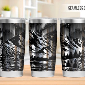 Puede incluir: Vaso de acero inoxidable con un diseño de montaña y aurora boreal en blanco y negro. El vaso tiene una tapa y base plateadas, y las palabras "SEAMLESS DESIGN" están impresas en la parte superior derecha.