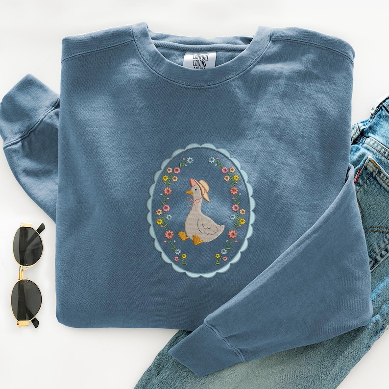 Puede incluir: Sudadera azul con cuello redondo y mangas largas. Presenta un dise&ntilde;o ovalado bordado con un ganso con sombrero, rodeado de flores coloridas. La sudadera se muestra con vaqueros azules y gafas de sol.