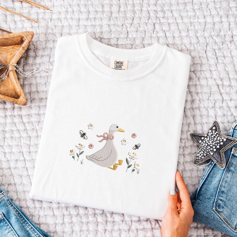 Op de afbeelding: Een wit Comfort Colors T-shirt met een geborduurd ontwerp van een grijze gans met een roze strik, omringd door bloemen en bijen. Het T-shirt is netjes op een getextureerd oppervlak gevouwen.