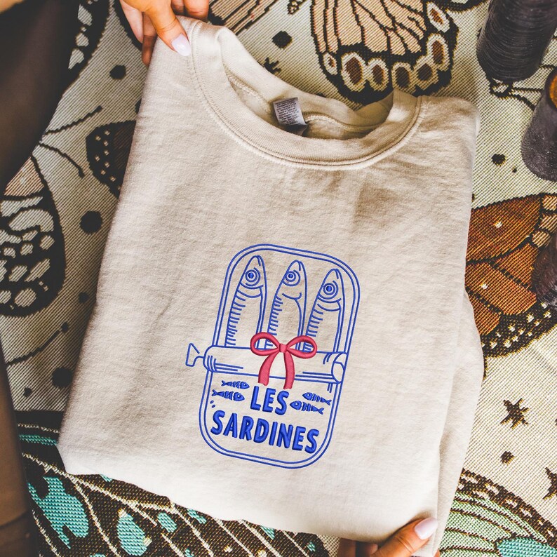 Peut inclure: Un sweat-shirt &agrave; col rond beige avec une broderie bleue repr&eacute;sentant trois sardines dans une bo&icirc;te de conserve ouverte. Le motif comprend un petit n&oelig;ud rouge nou&eacute; au centre de la bo&icirc;te et le texte LES SARDINES en lettres bleues.