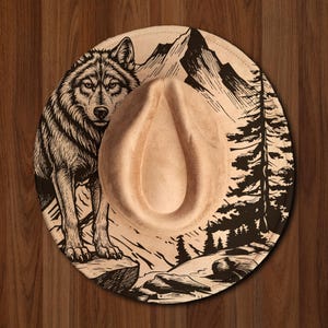 May include: A tan-colored fedora-style hat with a detailed illustration of a wolf, mountains, and trees. The hat has a teardrop-shaped crown and a wide brim. The artwork is in black and white.