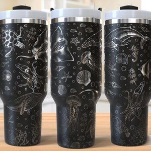Puede incluir: Tres tazas de viaje de acero inoxidable negro con un diseño plateado de varias criaturas marinas, incluyendo una ballena, una medusa, un caballito de mar y una estrella de mar. Las tazas tienen un asa negra y una tapa negra. El texto "SEAMLESS DESIGN" es visible en la parte inferior de la imagen.