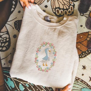 Puede incluir: Sudadera color crema con un dise&ntilde;o de ganso bordado dentro de una corona floral. El ganso es azul claro con pico y patas amarillas, con un lazo azul. La corona incluye flores rosas y azules y hojas verdes.